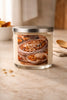 Clear glass jar candle with silver lid and pale cream wax; front label reads "Peach Cobbler" showing a cobbler image.