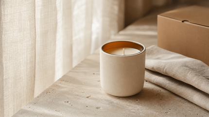 Unlit ivory candle beside linen and a shipping box in a shaded sunlit room