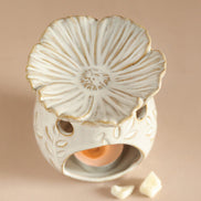 Cream ceramic warmer with gold-rimmed flower top, hollow base with leaf motifs and circular cutouts, lit tealight and two wax pieces on beige surface.