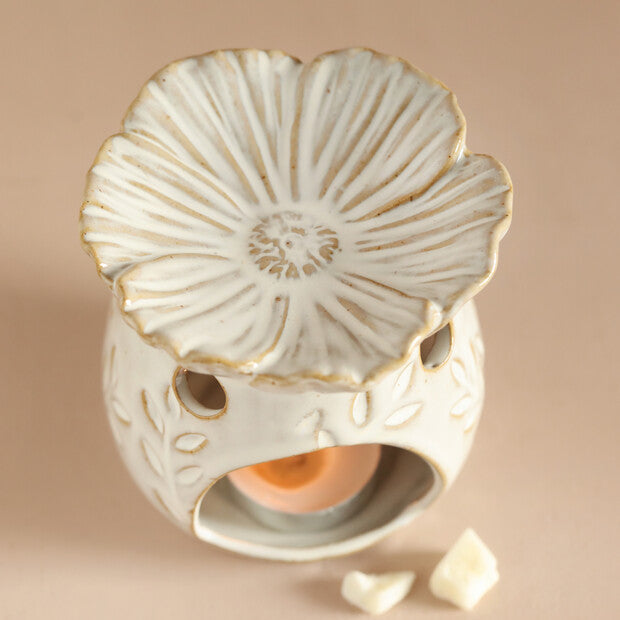 Cream ceramic warmer with gold-rimmed flower top, hollow base with leaf motifs and circular cutouts, lit tealight and two wax pieces on beige surface.