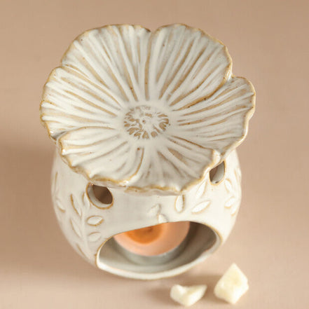 Cream ceramic warmer with gold-rimmed flower top, hollow base with leaf motifs and circular cutouts, lit tealight and two wax pieces on beige surface.