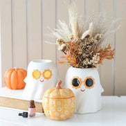 Two white ghost warmers with flower sunglasses (yellow-orange, black); right has dried beige-orange foliage, with orange pumpkin.