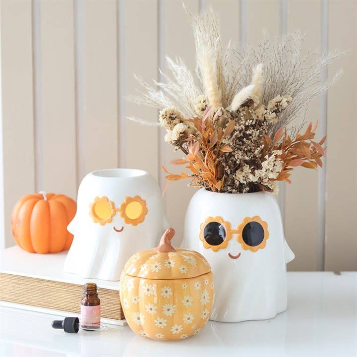 Two white ghost warmers with flower sunglasses (yellow-orange, black); right has dried beige-orange foliage, with orange pumpkin.