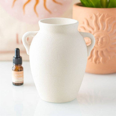 Matte off-white textured ceramic vase oil burner with two loop handles, flanked by an amber dropper bottle and terracotta sun planter.