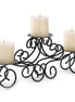 Black metal scrollwork candle centerpiece with three lit white pillar candles on staggered round trays.