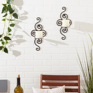 Two dark metal scrollwork wall sconces holding white pillar candles on a white brick wall above a patio table.