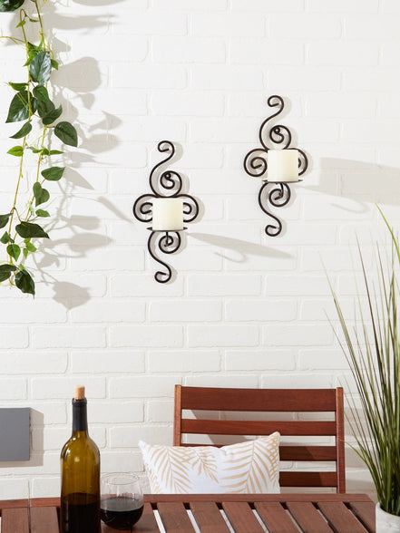 Two dark metal scrollwork wall sconces holding white pillar candles on a white brick wall above a patio table.