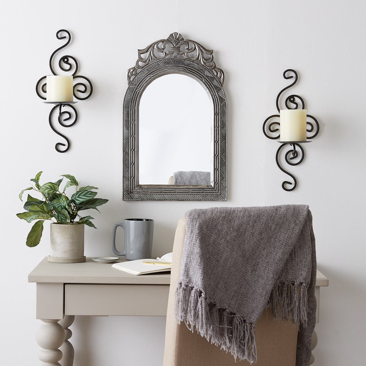 Pair of dark metal scrollwork wall sconces holding white pillar candles on either side of an arched mirror above a desk.