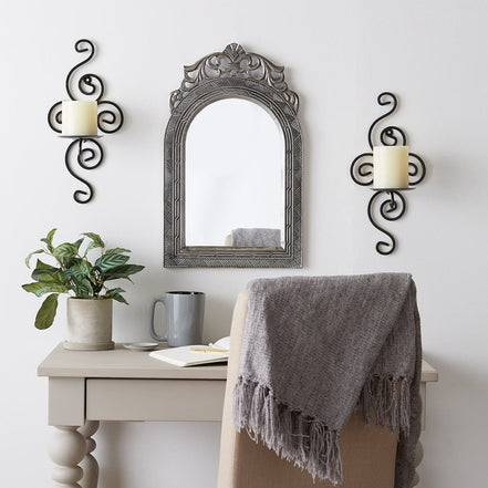 Pair of dark metal scrollwork wall sconces holding white pillar candles on either side of an arched mirror above a desk.