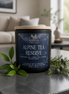 Alpine Tea Reserve candle in a dark glass jar with a wood lid, styled with evergreen sprigs in a light room.