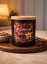 Black jar candle with wood lid labeled Phoenix Crafted Ambered Vanilla on a wooden tray with a ceramic bowl of amber beads.