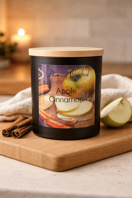 Apple Cinnamon candle in a black jar with wooden lid, styled with fresh green apple slices and cinnamon sticks on a wooden surface, all‑natural hand‑poured candle by Phoenix Crafted in a warm, cozy setting