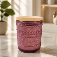 Purple hobnail glass candle jar with round light wood lid on a sunlit marble countertop, blurred plant and shelving in the background