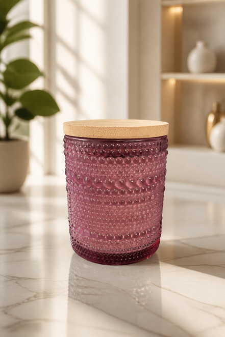 Purple hobnail glass candle jar with round light wood lid on a sunlit marble countertop, blurred plant and shelving in the background