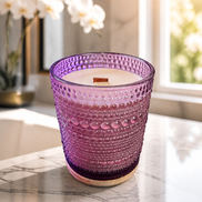Cream wax candle with a single wooden wick in a purple hobnail glass vessel on a round wooden coaster.
