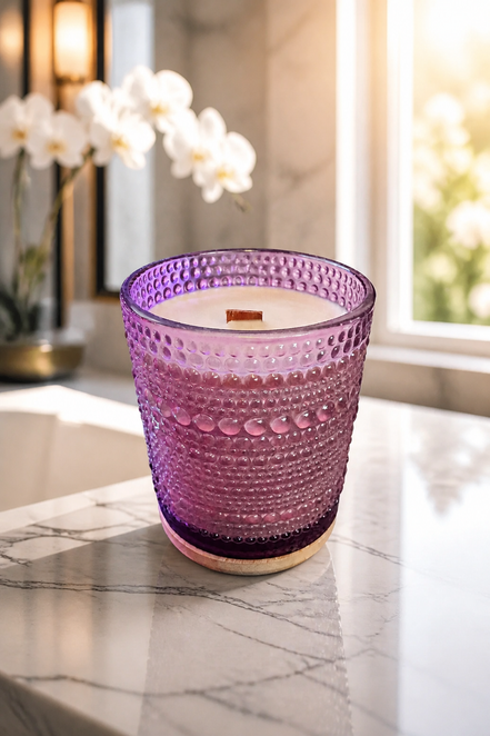 Cream wax candle with a single wooden wick in a purple hobnail glass vessel on a round wooden coaster.