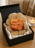 Pale beeswax tealights in clear cups, shrink-wrapped on cream cloth inside a matte black gift box on a wood table, one lit votive at right.