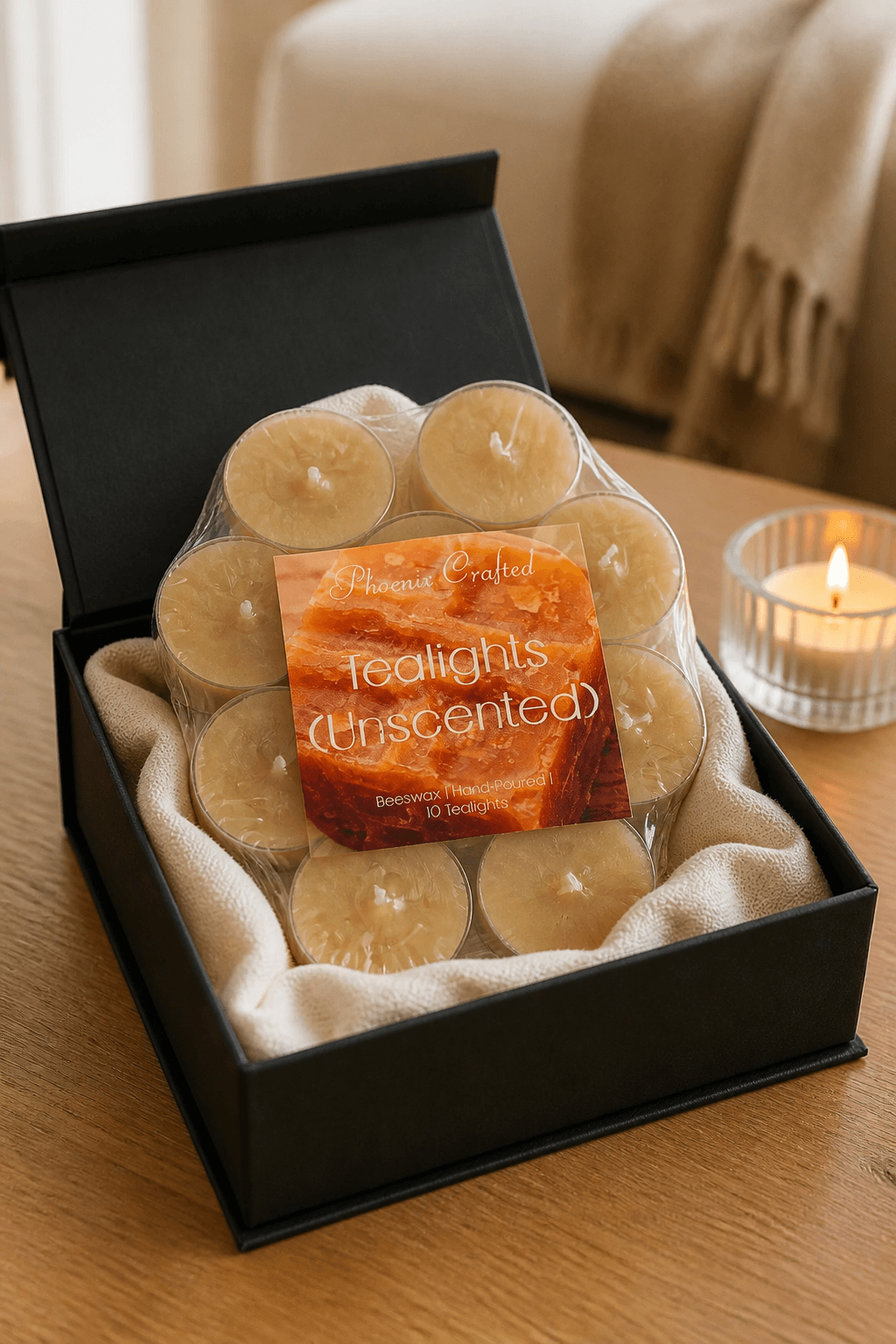 Pale beeswax tealights in clear cups, shrink-wrapped on cream cloth inside a matte black gift box on a wood table, one lit votive at right.