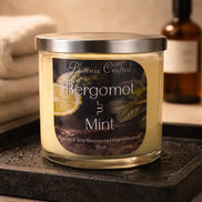 Phoenix Crafted Bergamot & Mint candle — pale yellow wax in clear glass jar with silver lid and black label, set on a wet black tray.