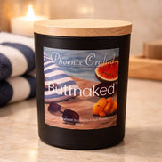 Black matte jar candle with light wood lid labeled 'Phoenix Crafted' and 'Buttnaked' beside a striped beach towel, sunglasses.