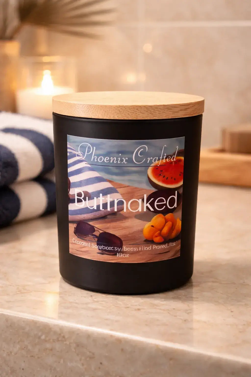 Black matte jar candle with light wood lid labeled 'Phoenix Crafted' and 'Buttnaked' beside a striped beach towel, sunglasses.