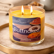 Yellow two-wick candle in a clear glass jar with a 'Phoenix Crafted' label reading 'Buttnaked' showing coconut and mango.