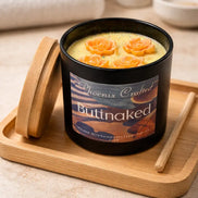 Black glass candle labeled 'Phoenix Crafted Buttnaked,' pale yellow wax topped with four orange rose decorations, short wick visible.