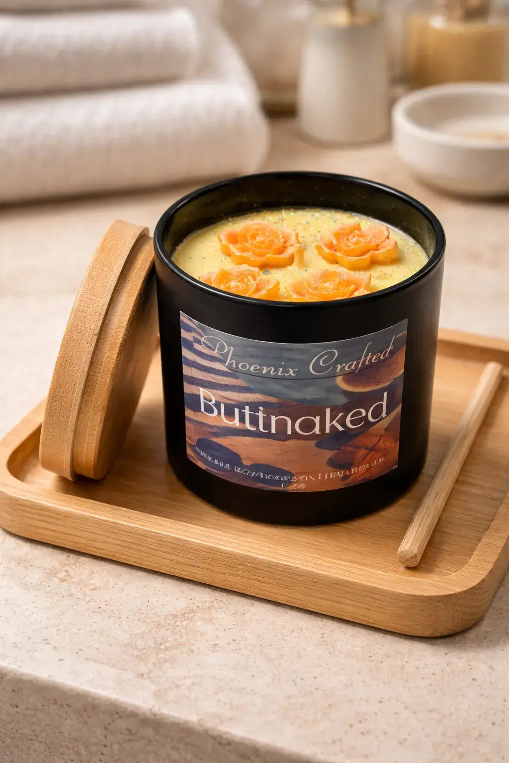 Black glass candle labeled 'Phoenix Crafted Buttnaked,' pale yellow wax topped with four orange rose decorations, short wick visible.