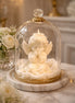 Caramel Apple decorative angel candle inside a clear glass cloche with gold trim, styled on a pale marble surface beside white flowers.