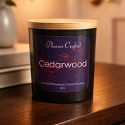Black matte jar candle with light wooden lid; purple label reads 'Phoenix Crafted' and 'Cedarwood' with '14oz', placed on a dark wood surface.
