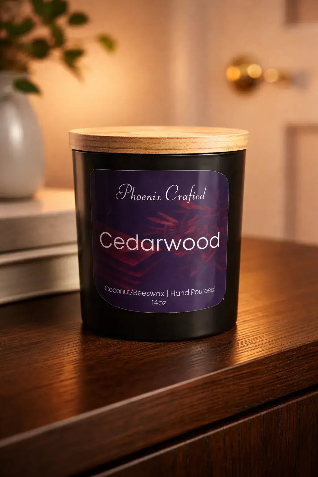 Black matte jar candle with light wooden lid; purple label reads 'Phoenix Crafted' and 'Cedarwood' with '14oz', placed on a dark wood surface.