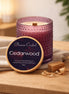 Textured purple glass candle with cream wax and visible wood wick, wooden lid propped showing the readable 'Cedarwood' label.