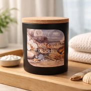 Coastal Orchid Woods Candle displayed on a wooden tray with seashells and a soft towel in a serene setting.