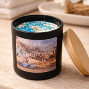 Matte black cylindrical candle labeled 'Coastal Orchid Woods' with white wax topped by blue crystals and a short wooden wick.