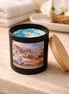 Matte black cylindrical candle labeled 'Coastal Orchid Woods' with white wax topped by blue crystals and a short wooden wick.