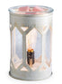 Cylindrical white distressed metal wax warmer with geometric cutouts and seeded glass panels; lit filament bulb inside.