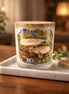 Frosted glass candle jar with round wooden lid labeled "Enchanted Forest" featuring a mushroom-forest photo, on a marble tray over a wood table.