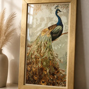 Gold-framed diamond-style peacock painting with a blue head and green-brown tail leaning on a stone tabletop beside a beige vase of pampas grass.
