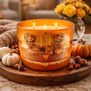 Three-wick amber glass Home Sweet Home candle on a wooden tray with small white and orange pumpkins and nuts.
