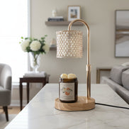 Brass arched lamp warmer with faceted glass shade and light wood oval base over an amber jar candle with orange label on a white marble table