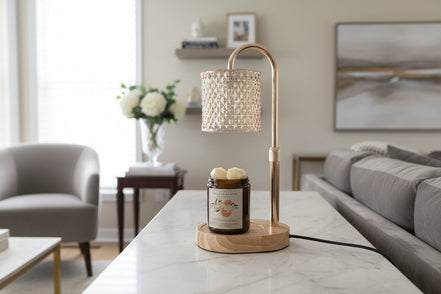 Brass arched lamp warmer with faceted glass shade and light wood oval base over an amber jar candle with orange label on a white marble table