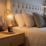 Lamp warmer with curved metal stem, textured beige drum shade and wooden base over an amber glass jar on an oak nightstand beside a tufted beige.