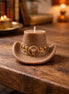 Leather cowboy-hat-shaped decorative candle with gold chain detail and a bull-head emblem, styled on a rustic wood table.