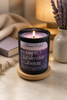 Lit Linen & Lavender Fields candle in a matte black jar on a wooden coaster beside lavender sprigs and a book.