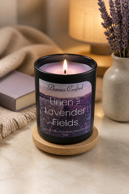 Lit Linen & Lavender Fields candle in a matte black jar on a wooden coaster beside lavender sprigs and a book.