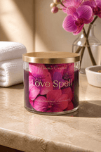 Candle labeled 'Love Spell' with a gold lid on a marble surface, surrounded by flowers.