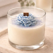 Clear round glass candle with white wax, blue layered flower topper and purple glitter, square wooden wick, on a beige stone tray