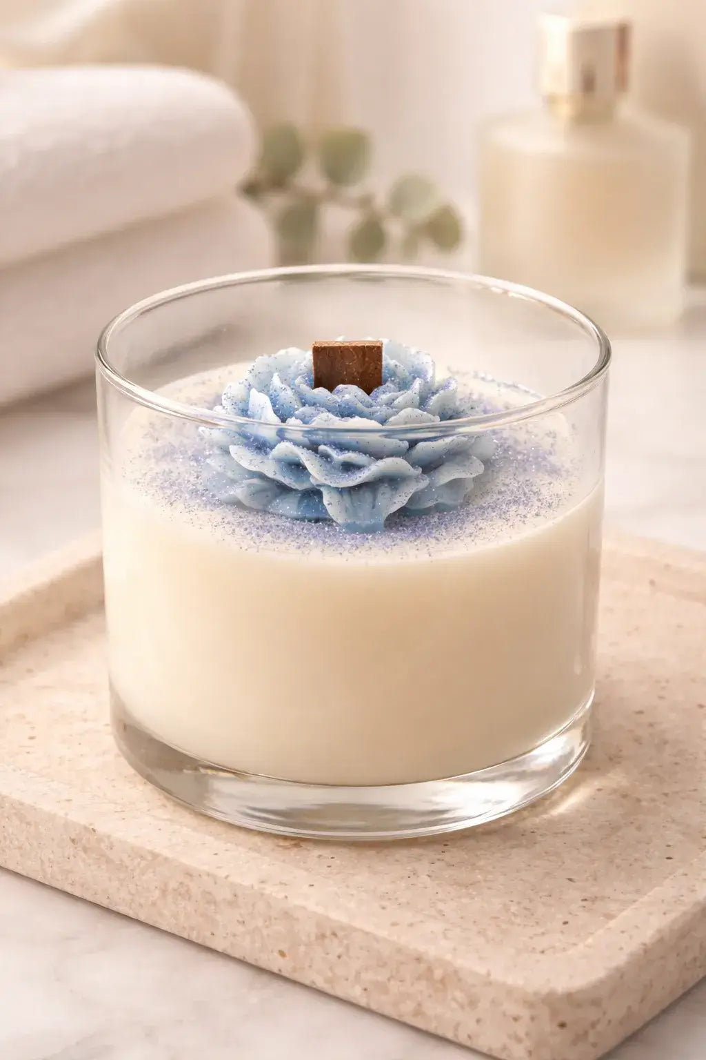 Clear round glass candle with white wax, blue layered flower topper and purple glitter, square wooden wick, on a beige stone tray