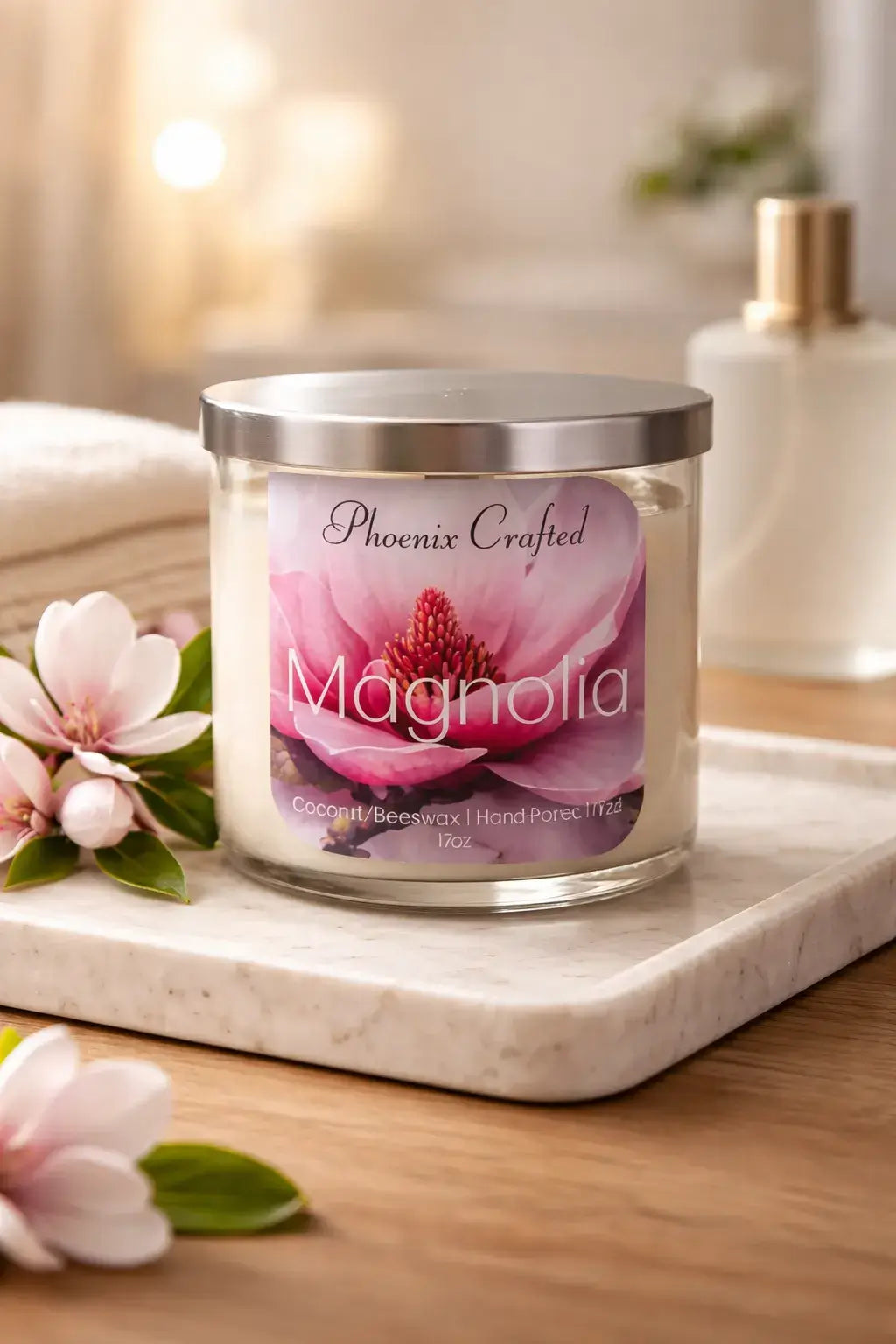 Clear glass jar candle with silver lid and Phoenix Crafted Magnolia label, white wax on a marble tray with pink magnolia blossoms.