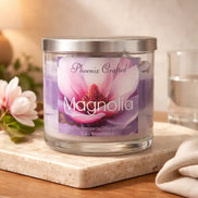 Clear glass jar candle with silver lid and purple layered wax; label reads Phoenix Crafted and Magnolia with magnolia artwork on a marble tile.